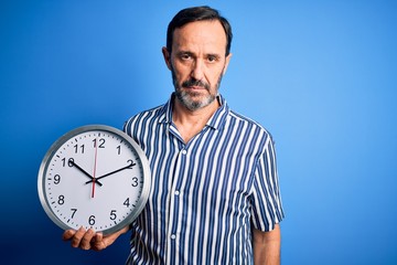 Middle age hoary man holding big clock standing over isolated blue background with a confident expression on smart face thinking serious