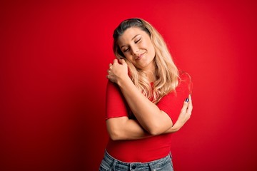 Young beautiful blonde woman wearing casual t-shirt standing over isolated red background Hugging...