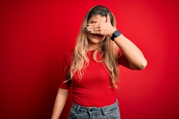 Young beautiful blonde woman wearing casual t-shirt standing over isolated red background covering eyes with hand, looking serious and sad. Sightless, hiding and rejection concept