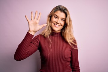Young beautiful blonde woman wearing casual sweater over isolated pink background showing and pointing up with fingers number five while smiling confident and happy.