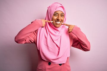 Young African American afro woman wearing muslim hijab over isolated pink background smiling cheerful showing and pointing with fingers teeth and mouth. Dental health concept.