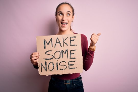 Beautiful activist woman holding banner with make some noise message over pink background pointing and showing with thumb up to the side with happy face smiling