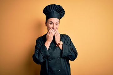Young beautiful chef woman wearing cooker uniform and hat standing over yellow background laughing...