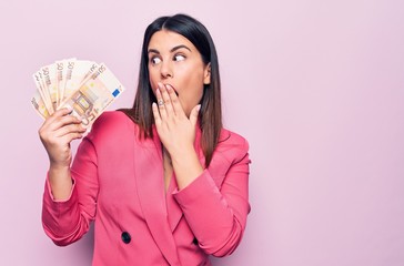 Young beautiful businesswoman holding euros banknotes over isolated pink background covering mouth...
