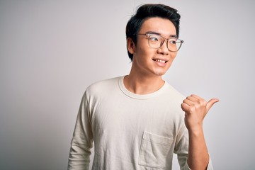 Young handsome chinese man wearing casual t-shirt and glasses over white background smiling with happy face looking and pointing to the side with thumb up.