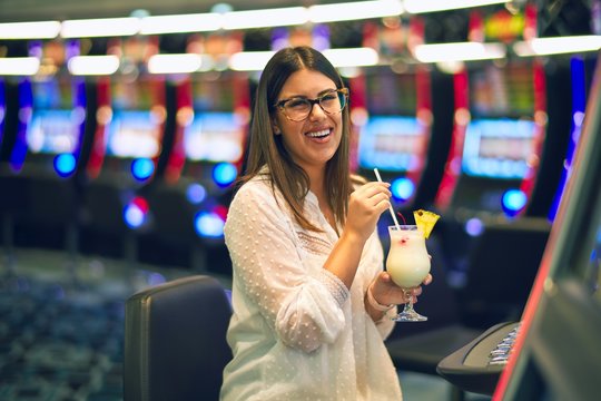 Young beautiful woman on vacation smiling happy and confident. Sitting with smile on face drinking cocktail and playing slot machine at casino