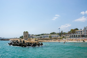 Seascape, view of a beach full with people, old tower of a castle and rocks and tetrapods, blue water and sky, summer season, nature