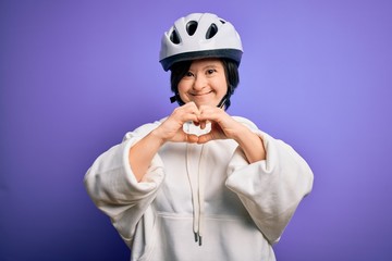 Young down syndrome cyclist woman wearing security bike helmet over purple background smiling in love showing heart symbol and shape with hands. Romantic concept.