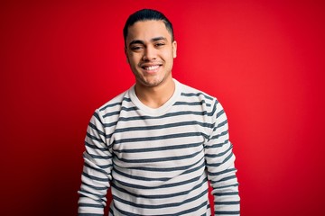 Young brazilian man wearing casual striped t-shirt standing over isolated red background with a happy and cool smile on face. Lucky person.