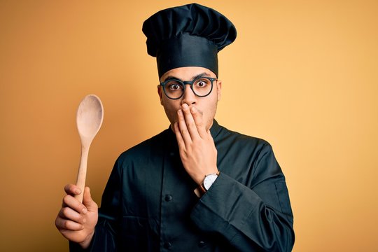 Young brazilian chef man wearing cooker uniform and hat holding wooden spoon cover mouth with hand shocked with shame for mistake, expression of fear, scared in silence, secret concept