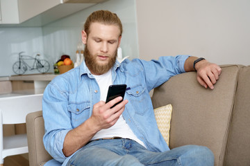 Fototapeta premium Serious young bearded man user holding smartphone texting message using online education app sit on sofa at home. Millennial guy using cellphone applications surfing web, read news on phone indoors.