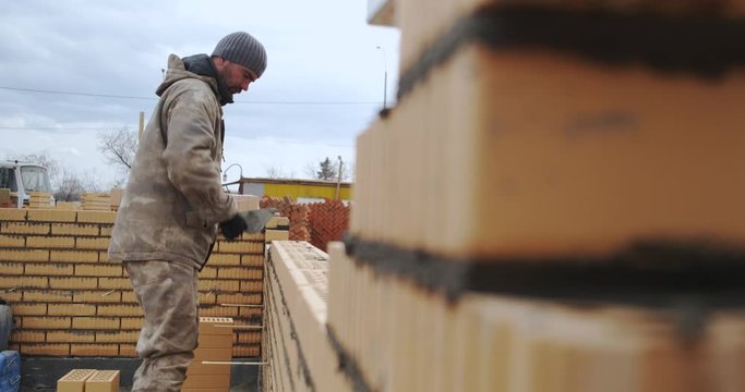 Male builder works at construction site as bricklayer, builds brick wall of warehouse, picks up brick, checks it, puts it in masonry, neat masonry, spring, construction season has begun, uniform