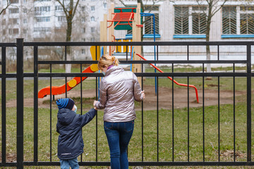 Mother and child in medical masks look at each other. On the background of a Playground behind a metal fence.