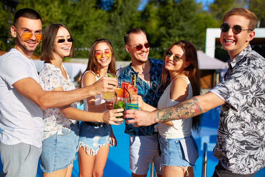 Group of friends having fun at poolside summer party clinking glasses with summer cocktails on sunny day near swimming pool. People toast drinking fresh juice at luxury villa on tropical vacation.