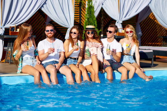 Group of friends having fun at poolside party clinking glasses with fresh cocktails sitting by swimming pool on sunny summer day. People toast drinking beverages at luxury villa on tropical vacation.