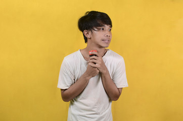 portrait of Asian man wearing a white shirt with a funny expression while carrying a flashlight isolated on a yellow background
