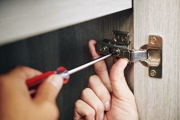 Close-up image of handyman assembling kitchen cabinet and screwing door hinge - obrazy, fototapety, plakaty