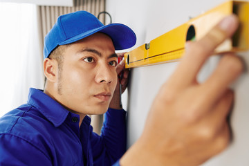 Serious young Asian repairman measuring wall with spirit level