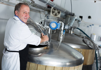 Adult brewer is standing with tube near reservoir for beer