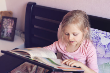 Beautiful girl sitting on the bed and reading a book. Looks at the camera, close-up. Distance Learning, Reading Learning, Quarantine Learning. Girl smiling and doing homework
