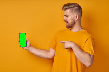 Happy Caucasian red haired man with thick beard, points at smart phone device, shows blank green screen with alfa channel for your promotional content, advertizes new device for customers, on orange