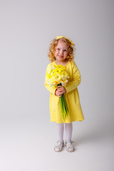 a small curly haired girl in a yellow dress holding a bouquet of spring flowers on a white background smiles