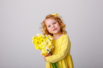a small curly haired girl in a yellow dress holding a bouquet of spring flowers on a white background smiles