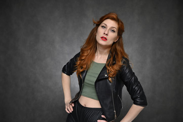 Obraz na płótnie Canvas Horizontal portrait of a red-haired young woman in a black jacket and green t-shirt. Model with excellent makeup posing on a gray background in the studio.