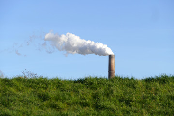 Rauchender Schornstein einer Fabrik hinter einem Deich am Rhein