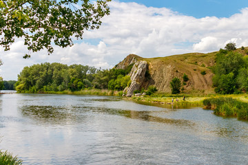 Fototapeta na wymiar An idyllic landscape view with a big rock on a riverbank and small figures of resting people on the water side