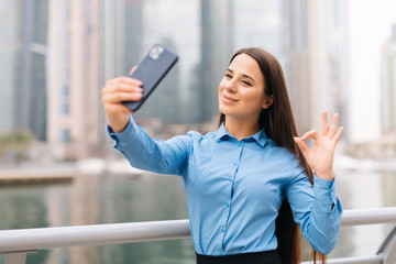 Fototapeta premium Happy young business woman make video call smart phone with okay gesture on broadwalk on skyscrappers background