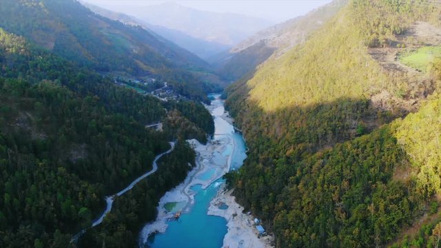 Drone shot of flying between a tight green valley river Tama koshi flowing between them during golden hour with road on the side. Aerial view on the way to Jiri, Dolakha. Developing nation Nepal 2019.