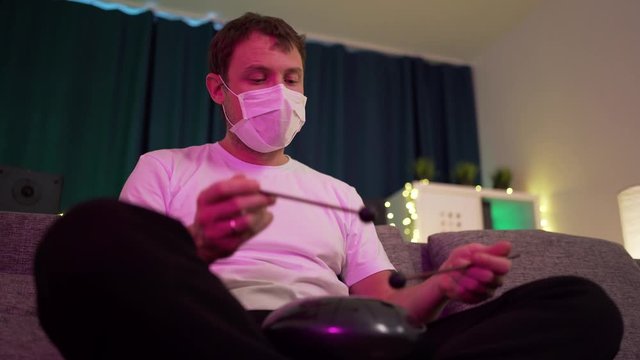 a man in a protective mask sits at home and plays on the hapi drum