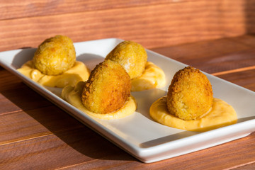 Traditional spanish snack- croquettes fried in oil, with yellow cream sauce on white plate at sunny kitchen. Side view on wooden background