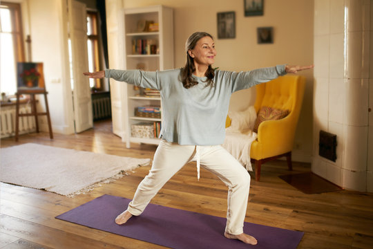 Full length shot of happy energetic mature woman in casual clothes exercising at home because of social distancing, practicing yoga on mat, standing in warrior ii pose. Age, wellness and health