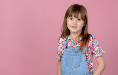 Sweet little girl 6-7 years old posing in dungarees jeans and flower pattern blouse on pink