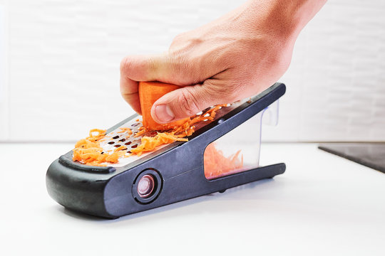 Man hand grating piece of carrot with steel grater on the white board in a kitchen.