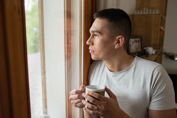 Casual young man at home holding a cup of coffee and looking out the window with his image reflected on the glass.