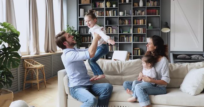Loving happy young parents playing with two cute small daughters in living room enjoy weekend activity. Cheerful family of four having fun together spending leisure free time at home sitting on couch.