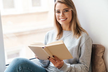 Photo of smiling woman reading book while sitting near window