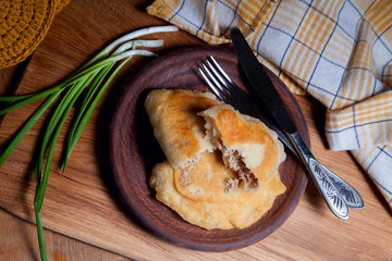 Clay plate of fried meat pies with cutlery and green onion on wooden table.