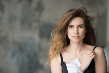 Obraz premium Beautiful elegant woman with flying hair and full lips in a white shirt on a gray background looks languidly at the camera. Beauty and fashionable style. Soft selective focus.