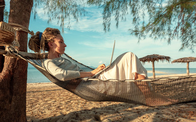 Young woman working on laptop lying in hammock at sand beach of tropical island. Freelance outdoor work concept