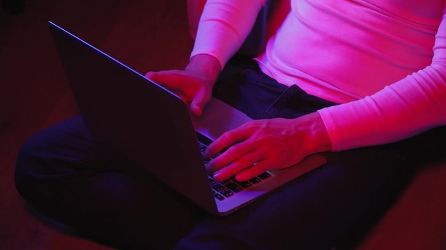 A young, sporty man prints on a laptop, at home. Close-up of hands of a guy in a white turtleneck, working remotely. The businessman prints on the computer, muffled light, twilight.