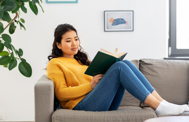 Fototapeta na wymiar people and leisure concept - happy asian young woman in yellow sweater sitting on sofa and reading book at home