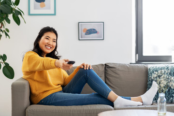 people and leisure concept - happy smiling asian young woman with tv remote control sitting on sofa at home