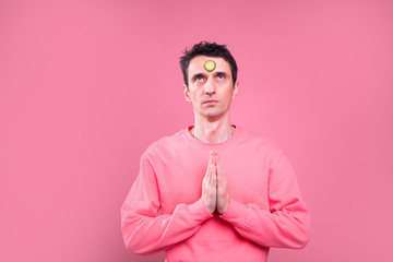 Serious concentrated young man stand alone and hold hands in praying position. One cucumber slice is on forehead. Isolated over pink background.