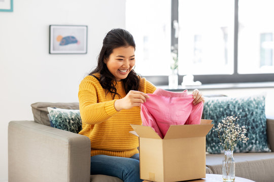 delivery, shipping and people concept - happy asian young woman taking clothes out of cardboard box or parcel at home