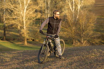 Sports brutal bearded guy on a modern mountain bike. Cyclist on the green hills in the spring.