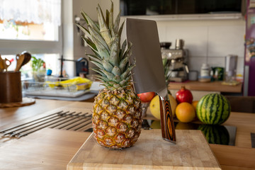 A cutting board with a whole pineapple fruit and a cleaver standing on a worktop in the kitchen in the background are more fruits and kitchen utensils
